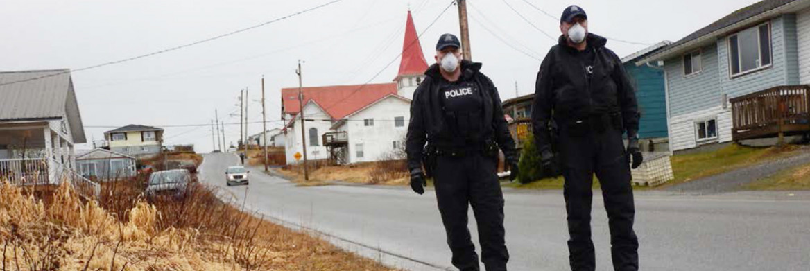 Two male RCMP officers walk on the side of a road wearing protective masks.