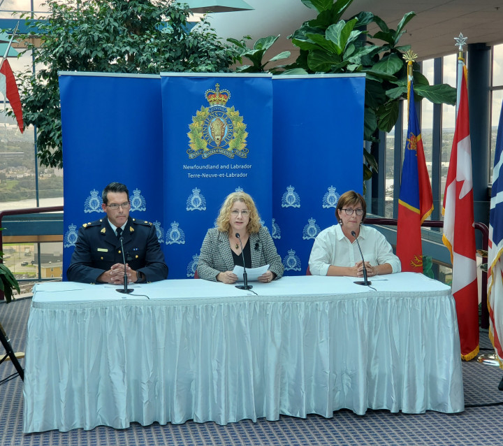 A man in a police uniform and two women sit at a table with microphones in front of them with an RCMP backdrop.