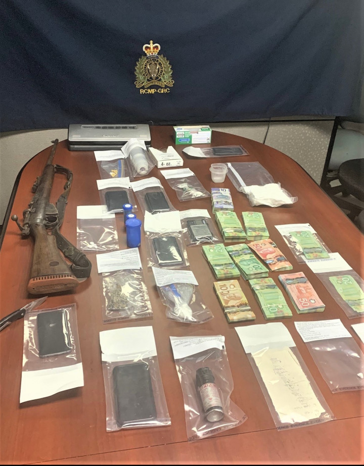 A large quantity of cash in sealed plastic bags is shown on a table, along with a brown rifle, cell phones, a vacuum sealer, scales, a knife and various bags of illicit drugs. 