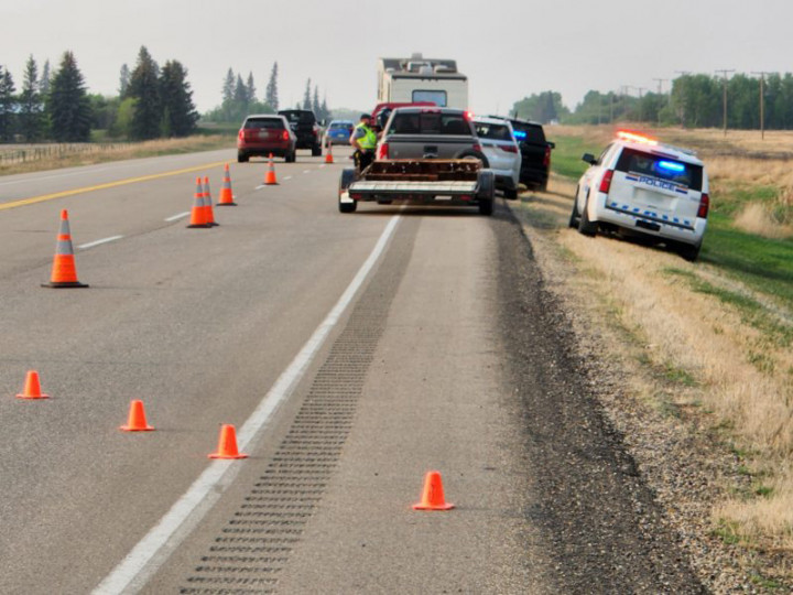 RCMP officer in fluorescent vest standing beside the window of a stopped grey truck pulling an empty trailer on the highway. RCMP vehicle with lights on in the right ditch beside the truck. Orange cones sectioning off the Highway in an 