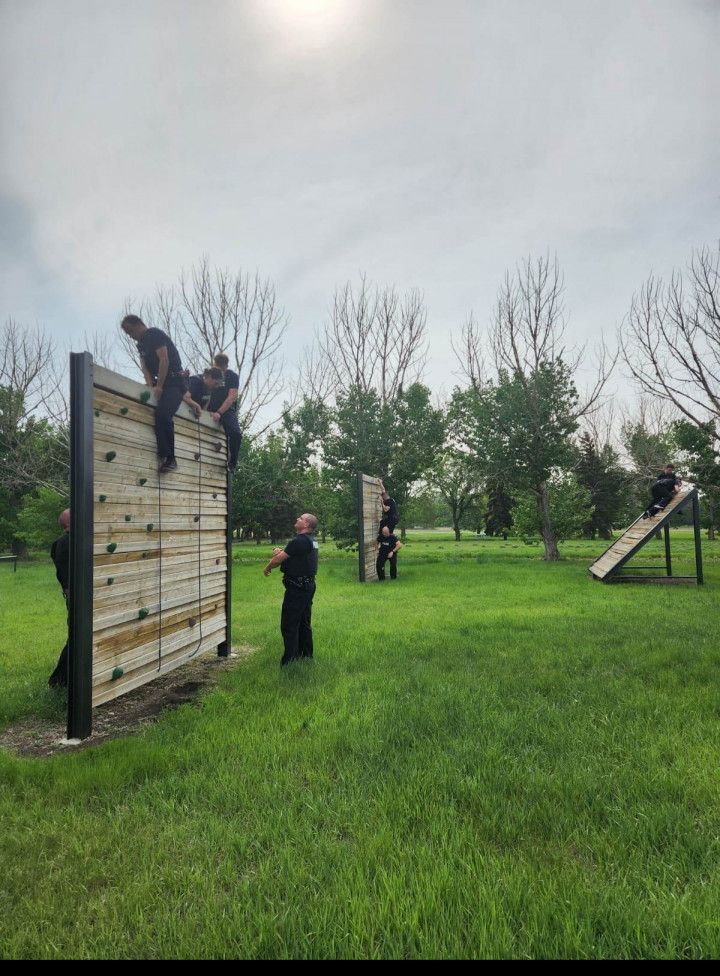 Groups of cadets wall climbing a wooden wall obstacle while two people supervise.