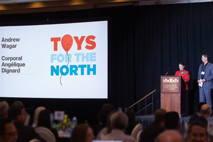 RCMP Cpl. Angelique Dignard and Andrew Wagar (Canadian Toys Association) on stage, in front a podium, to address guests at the Hall of Fame reception and dinner. Toys for the North logo is projected on a screen to the right of them. Part of the audience can be seen facing the stage.