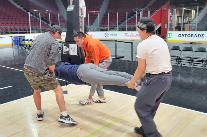 Inuit games airplane competition. In an arena, three people balance a 4th person who is stretched out horizontally in the air.