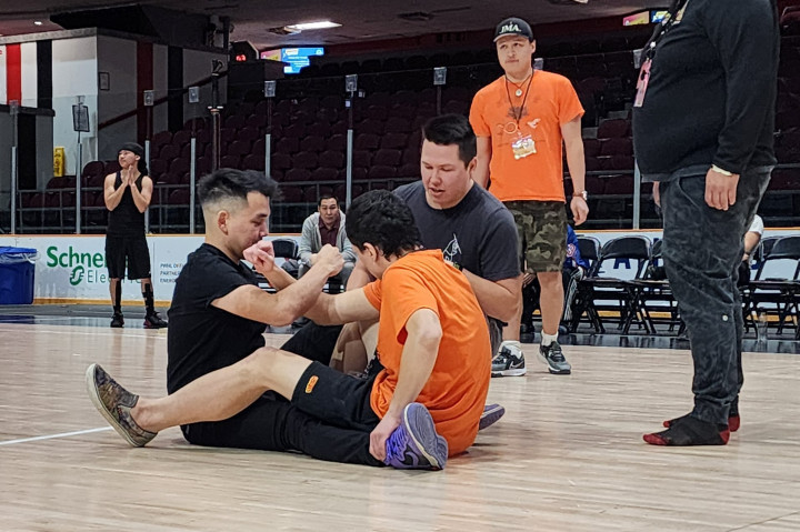 Inuit games arm pull competition. In an arena, two people are sitting on the floor, facing one another with an arm interlocked. Three other people are standing to supervise the competition. 