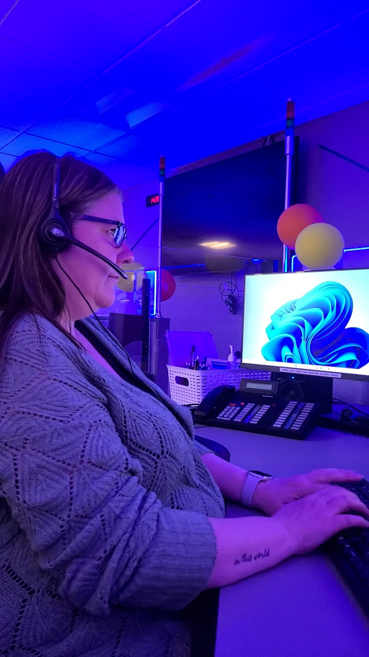 A woman wearing glasses, a grey sweater with a headset, typing on a computer 