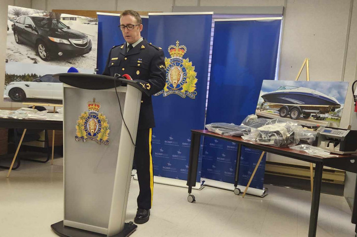 A uniformed police officer stands behind a podium with pictures of vehicles and a boat displayed on easels with blue RCMP banners in the background.