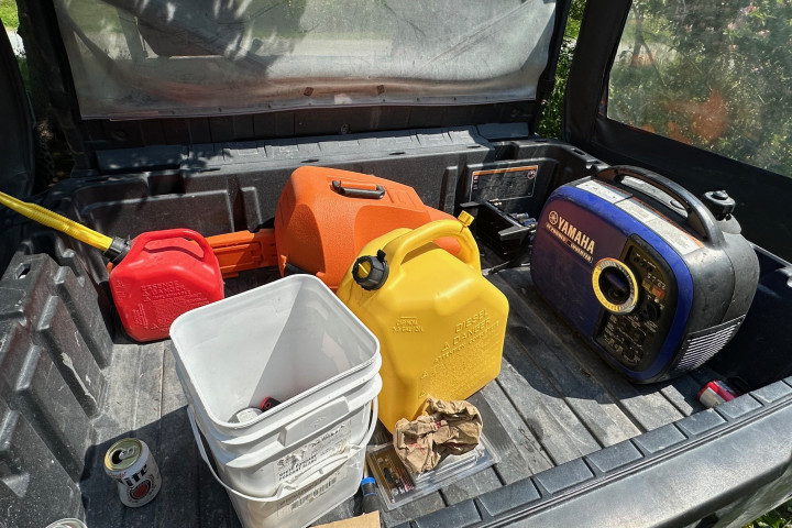 A generator, a chainsaw in an orange case, and some plastic fuel cans are pictured resting on the back of a UTV (side by side). 