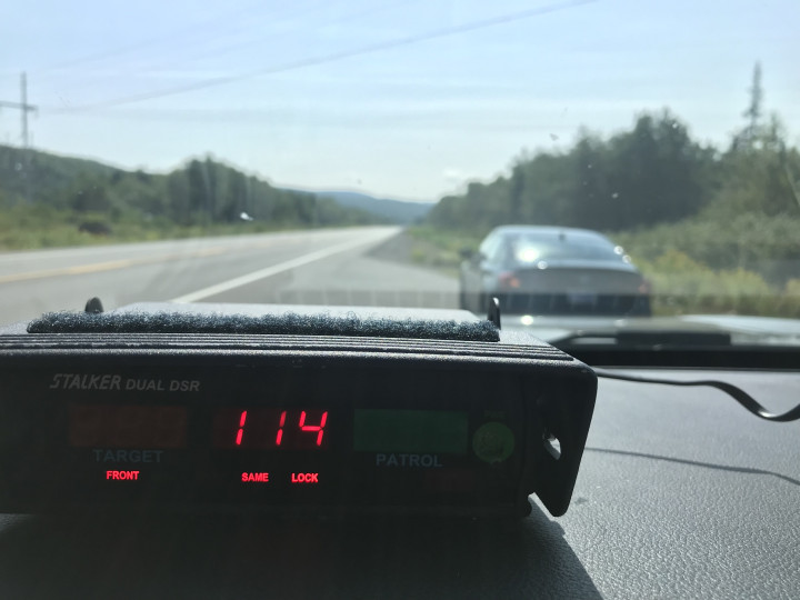 Un radar sur le tableau de bord d'un véhicule de police affiche une vitesse de 114 km/h. Une voiture noire est immobilisée sur le bord de la route devant le véhicule de police.