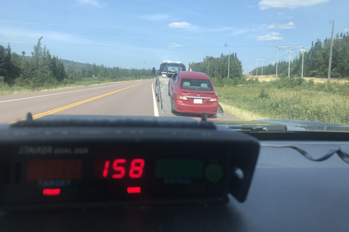 Un radar sur le tableau de bord d'un véhicule de police affiche une vitesse de 158 km/h. Une voiture rouge est sur le bord de la route devant le véhicule de police.