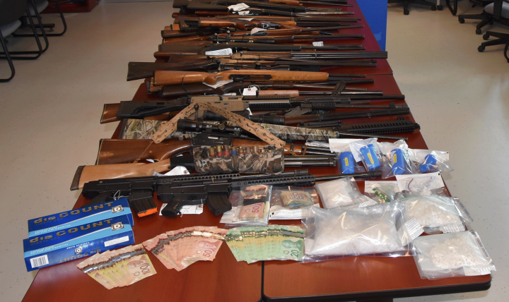 Rifles and shotguns displayed on a table with cash and clear bags of white powder. Two banners, one of Amherst Police Department and the other of Cumberland RCMP are in the background.