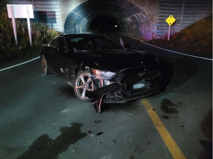 A damaged ford Mustang is parked on a roadway near the underpass of a highway.