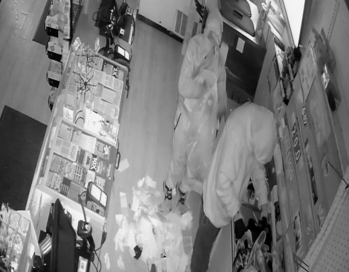 A still image taken from video surveillance footage, which shows two unknown suspects behind the cash register, with a pile of items on the floor. One suspect is seen handling items on the back counter, while the second stands and looks in the direction of the surveillance camera.