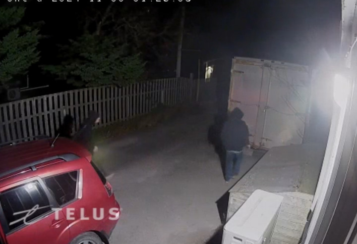 Two suspects wearing black hoodies are seen near a storage container outside alongside a fence at night. The back of a red or burgundy SUV can also be seen.