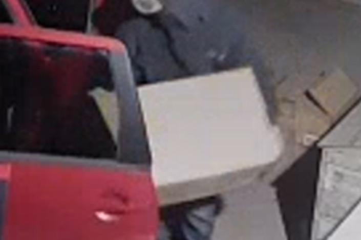 Suspect wearing a black hoodie over a baseball cap is putting a box into a red vehicle.