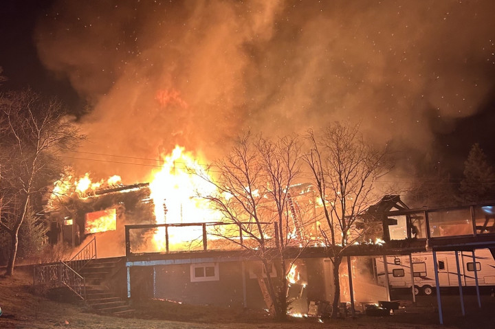 A cabin and trees on fire at night. There is a camper trailer in the background.