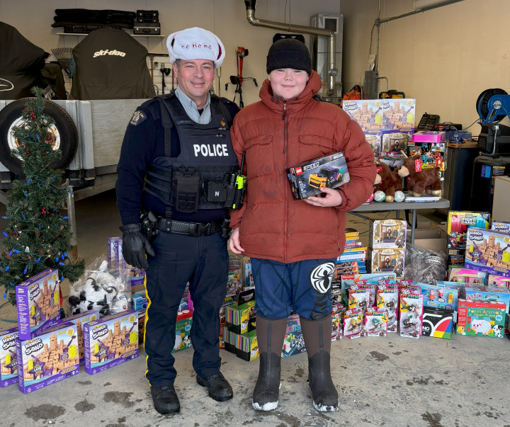 Un agent de la GRC en uniforme portant un bonnet de Père Noël se tient à côté d'un jeune tenant un jouet. Des jouets sont empilés derrière eux sur une table et sur le sol. 