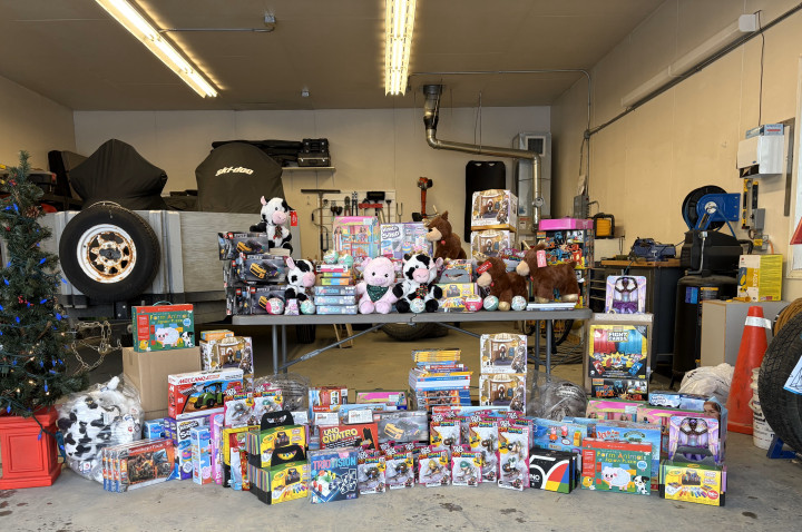 Des jouets non emballés sont empilés sur le sol et sur une table dans un garage. Il y a un arbre de Noël à côté de la table. 